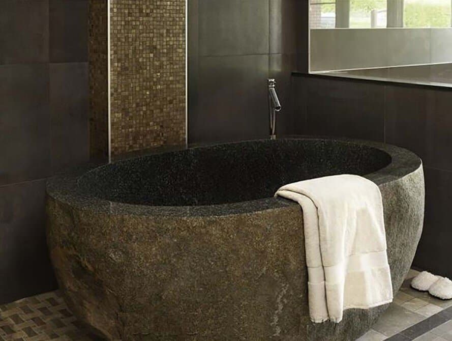 Carved dark stone freestanding bathtub in a spa-style bathroom with a mosaic tile accent wall
